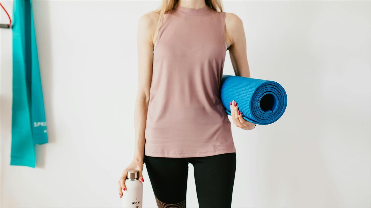 Dieses Bild zeigt eine Frau in einem ärmellosen rosa Oberteil und schwarzen Leggings, die eine zusammengerollte blaue Yogamatte in der einen Hand und eine weiße Wasserflasche mit der Aufschrift „I woke up“ in der anderen hält. Im Hintergrund hängt ein türkisfarbenes Fitnessband vor einer weißen Wand und vermittelt eine sportliche Atmosphäre.